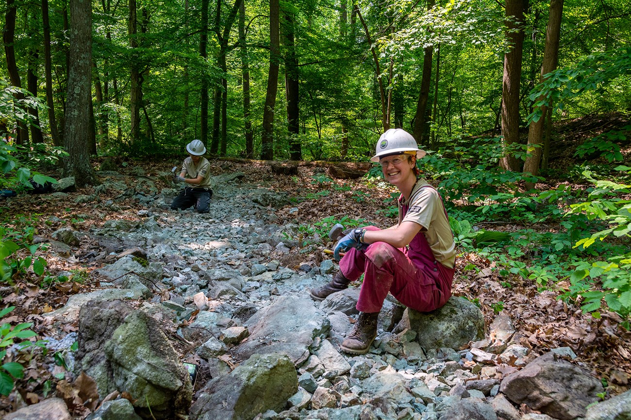 A young woman wearing brown pants and red pants sits on a rock among a field of rocks. Another individual sits in the back sitting on rocks