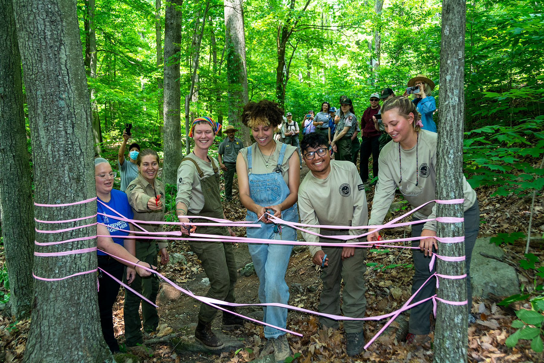 A group of six youth wearing shirts that say Conservation Legacy and SCA stand in the woods surrounded by other individuals. They are cutting a pink ribbon connected between two trees with scissors.