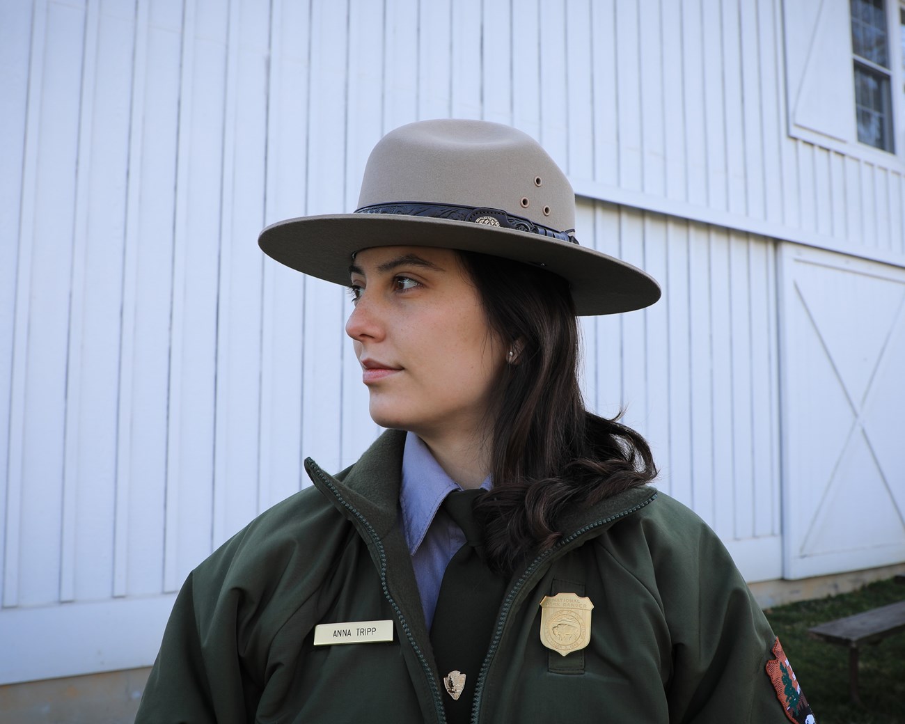 A white female National Park Service ranger poses looking off to the left in front of a white barn, part of a picnic table can be seen to her right.