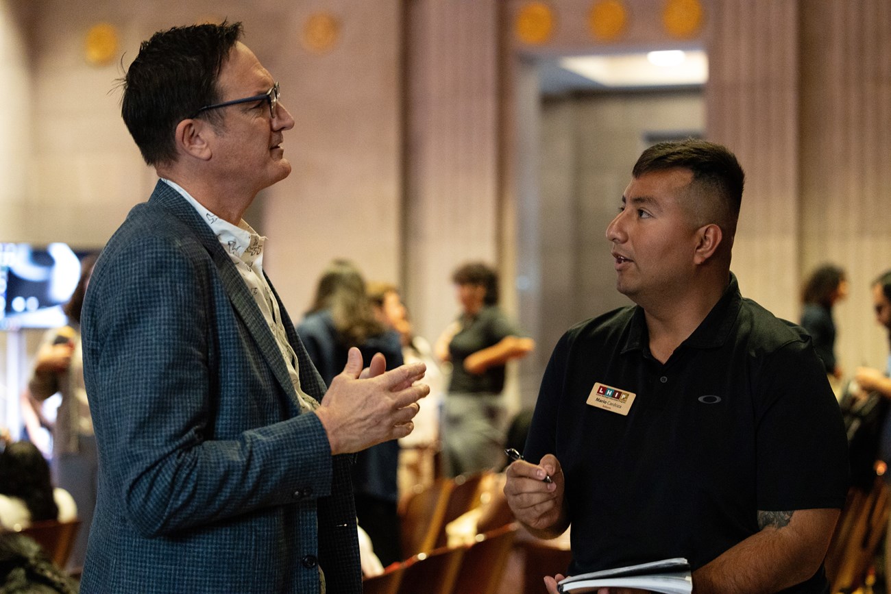 Two men, one wearing a suit, the other wearing a polo, stand facing each other in conversation