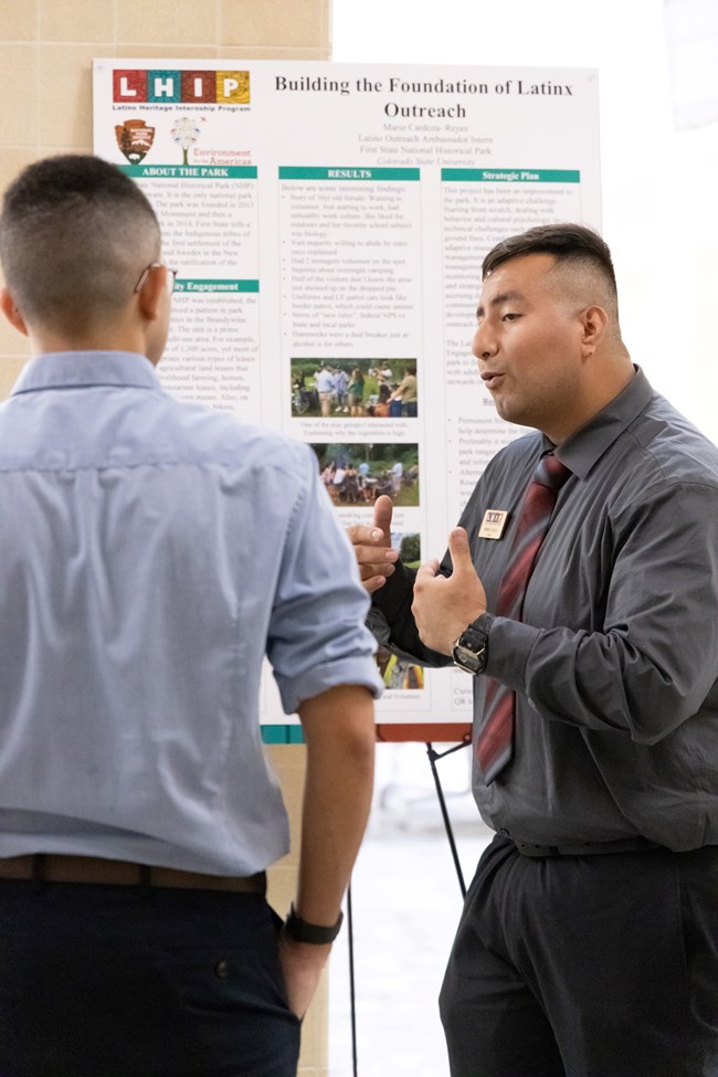 Two young men face each other. One is wearing professional attire and using hand gestures in front of a diagram