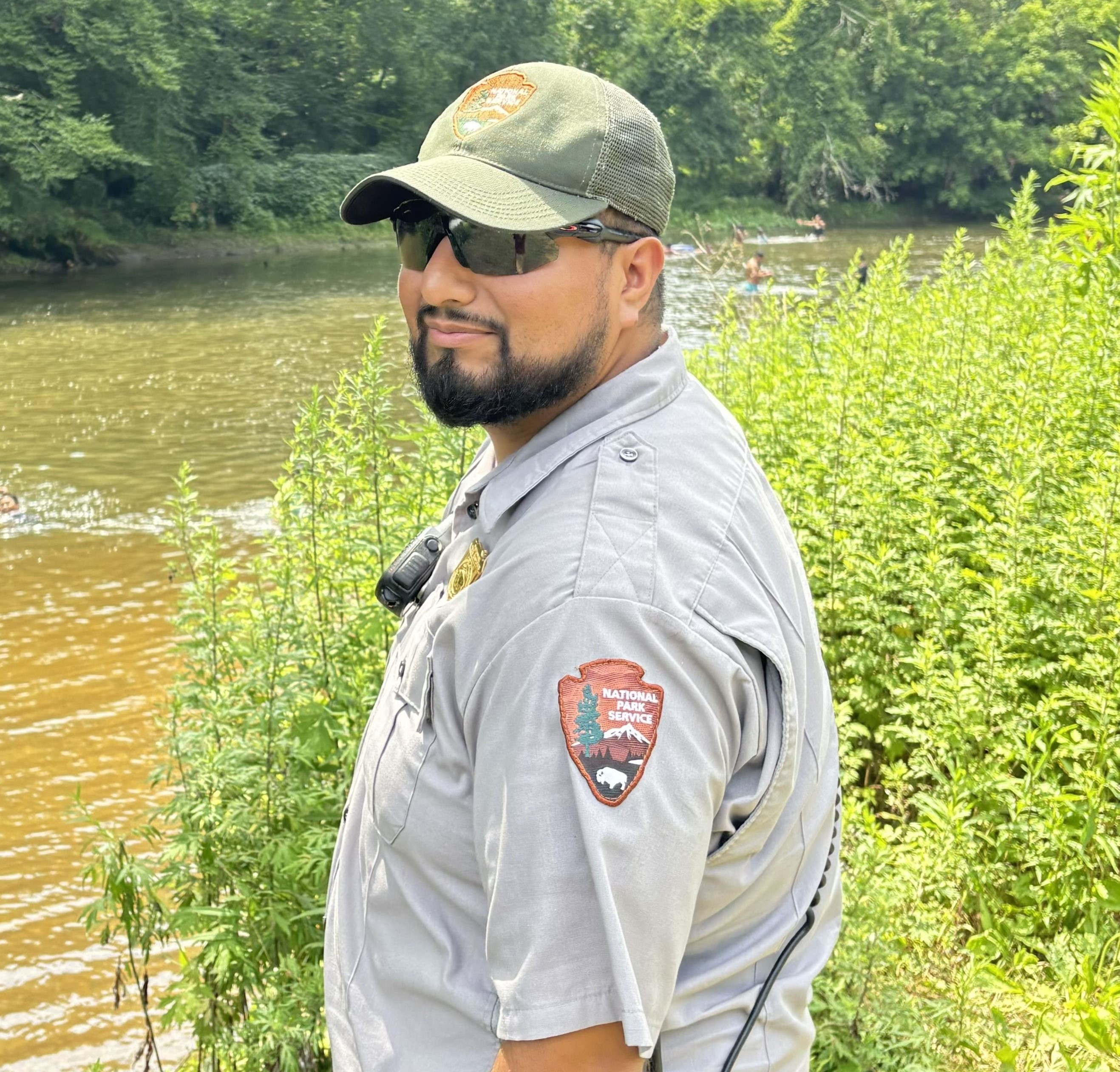 A man stands in front of a body of water wearing an NPS uniform and black sunglasses