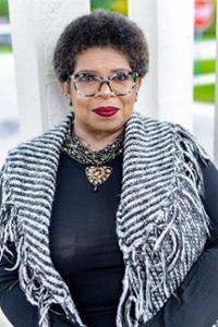 Black woman dressed in black with black and white striped shawl, tortoise shell glasses, and red lipstick.