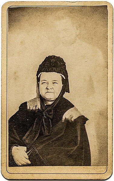 Mary Todd Lincoln, wearing a black dress and black bonnet, sits with her hands crossed. A ghostly figure with a beard appears to rest his hands on her shoulders.