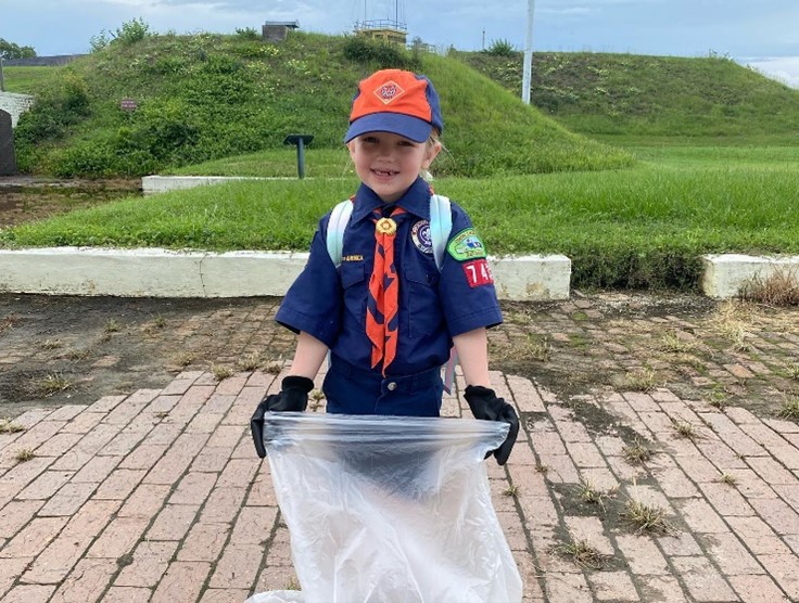 A young girl wearing a blue cub scout outfit and an orange hat holds a trash bag