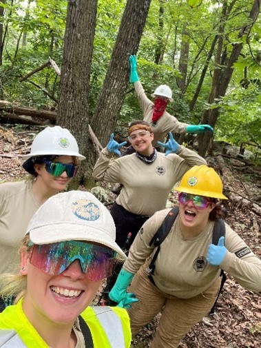 A group of five individuals wearing hard hats pose stand outside in front of trees. Three are holding up their hands with their thumbs ups.