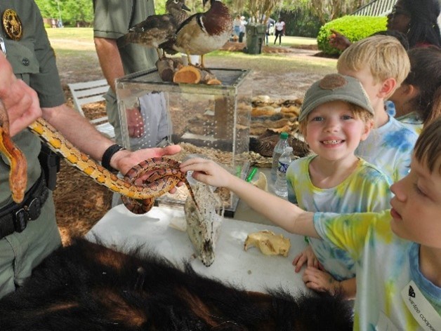 A young girl stands with two other youth in tye-die shirts. They are standing at a table petting snakes.