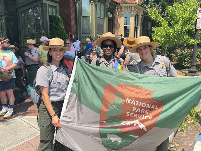 Three female National Park Service rangers hold NPS flag on a busy street