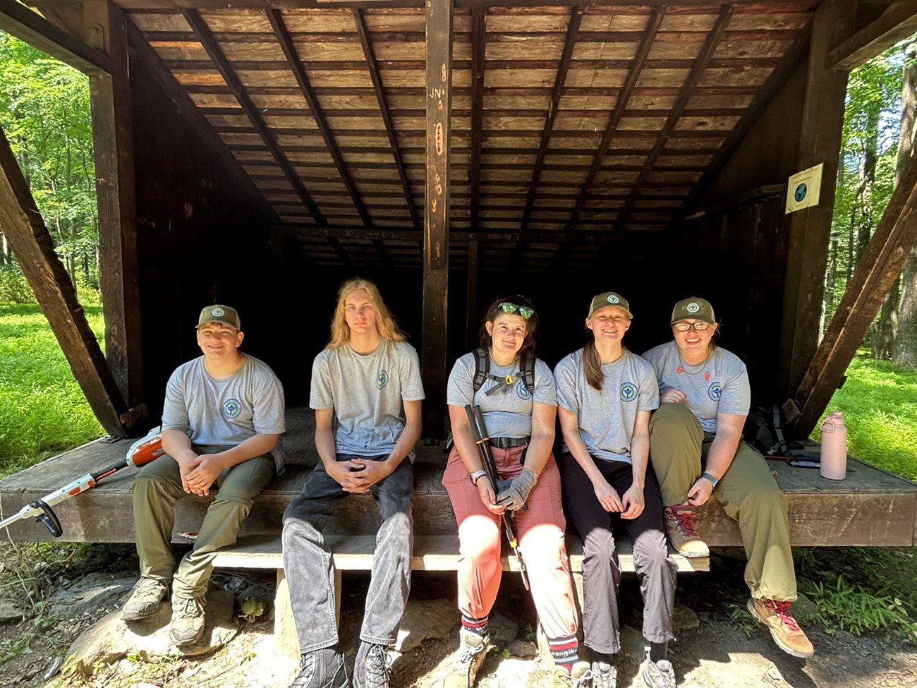 Five youth wearing hats sit outside under a shed structure. There are two mowing devices next to them.