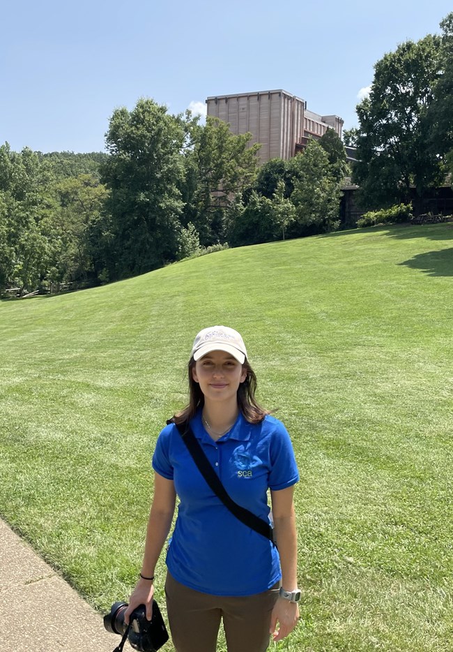 A white female stands in front of an open grassy area, with a wooden amphitheater behind trees and a camera in her hand. She’s wearing a blue polo with a logo that reads “SCA”.