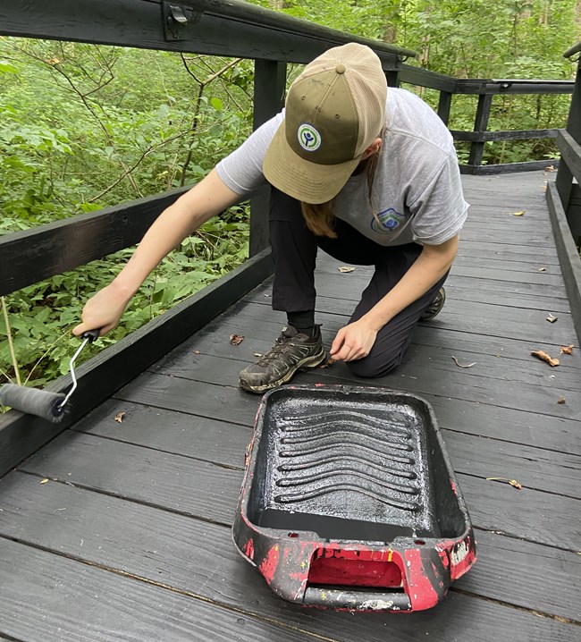 A young woman wearing a green ball cap kneels on a gray wooden bridge while painting the hand rail
