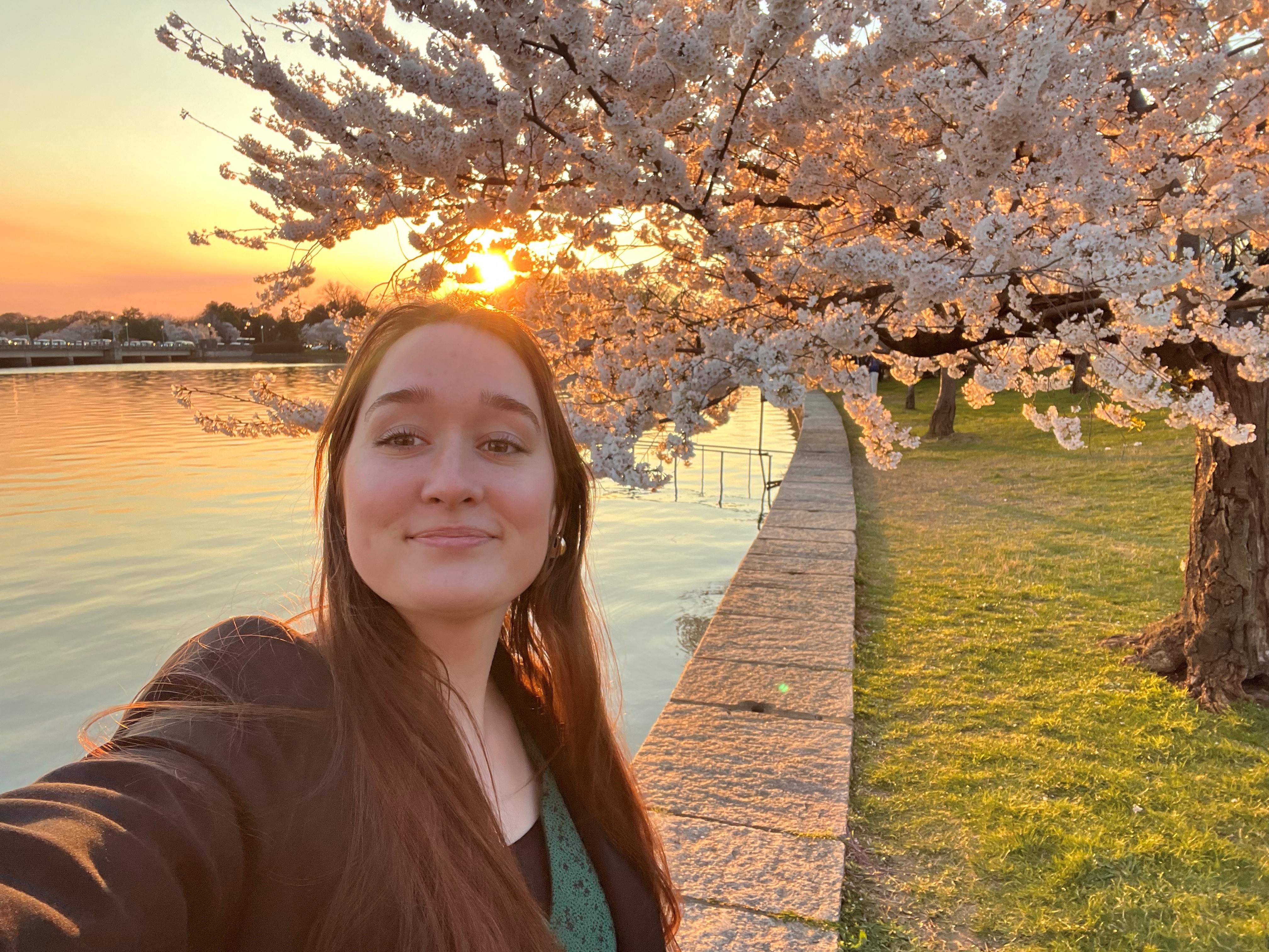 A young woman stands in front of a body of water smiling in front of a tree with cherry blossoms