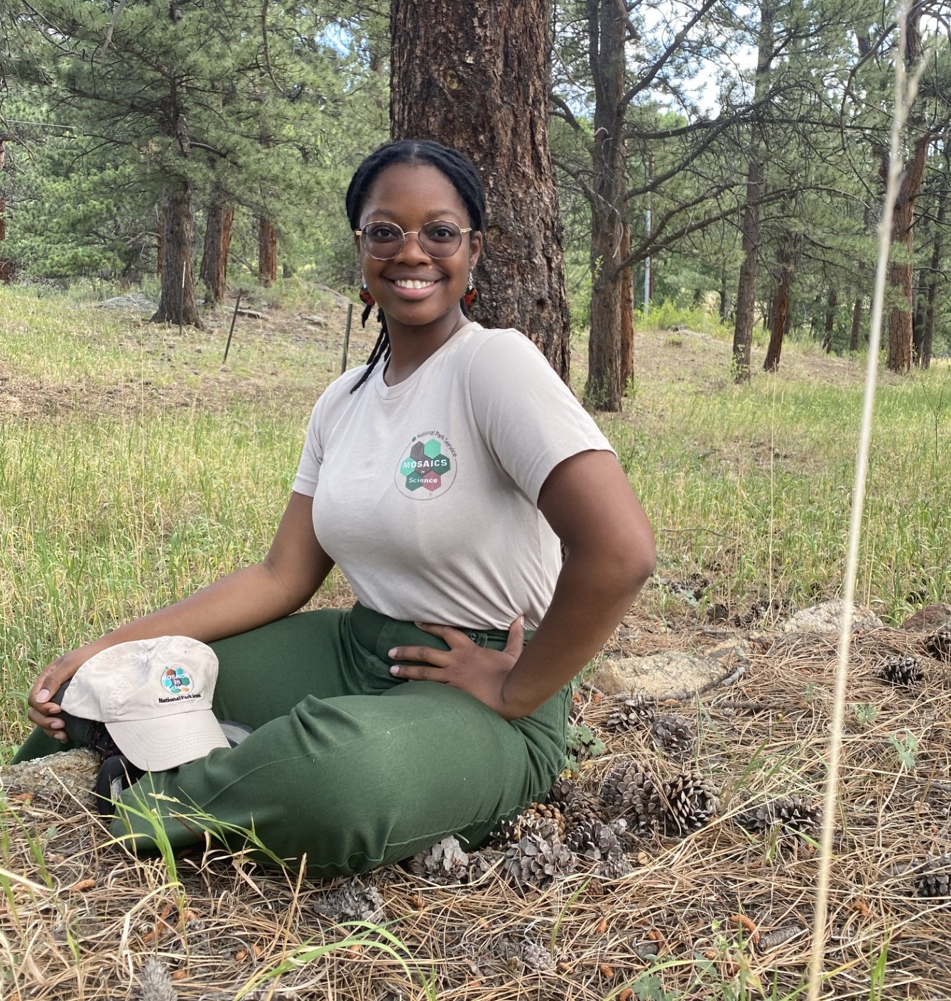 A young woman wearing a tan shirt with a logo that reads "Mosaics in Science" sits under a tree