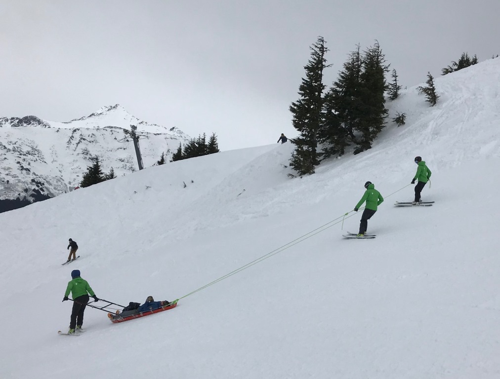 Three rangers carefully transport a patient in a sled down a ski slope.