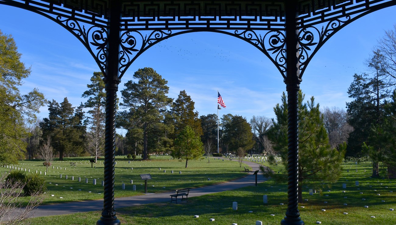 Ornate iron works frame a panorama of a cemetery with green trees, grass, white headstones, and a US flag flying in the center.