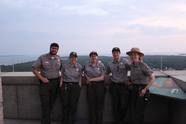 5 people wearing the national Park service uniform stand on the observation deck of the memotial with Lake Erie in the background.