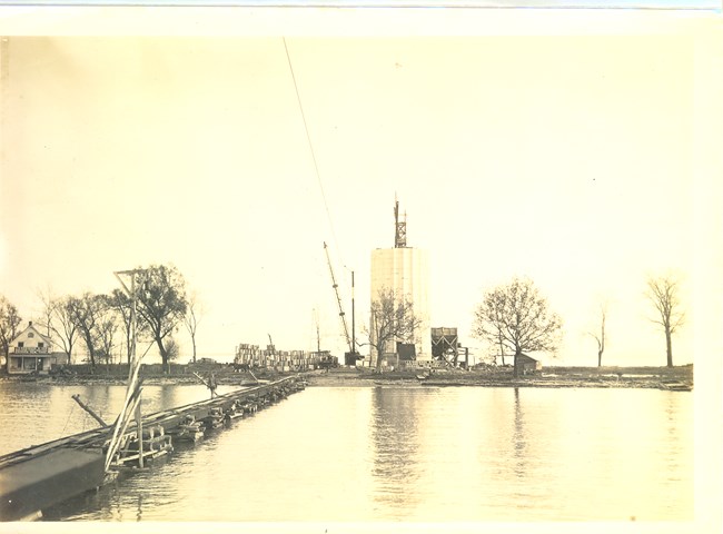 Monochrome photo from approximately 1913 with a 600-foot dock with rails built across a shallow bay to the island where a columnar monument is partially built.