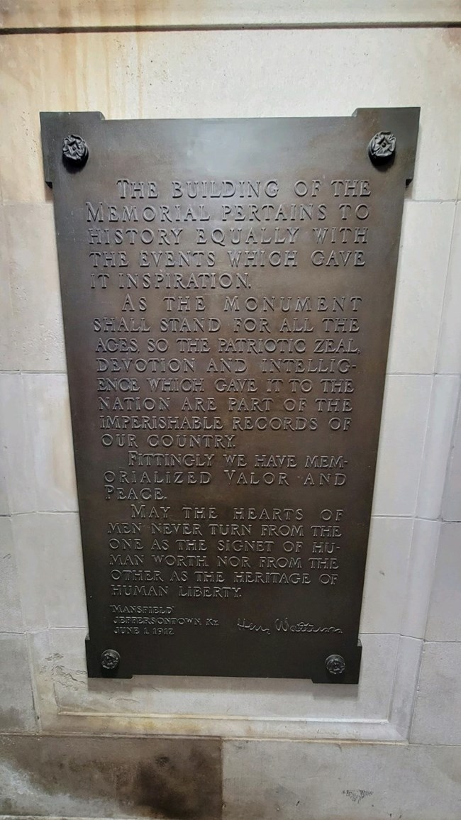 A bronze tablet hanging on the wall of a marble doorway. it is inscribed with a dedication memorializing patriotic zeal, valor, and more.