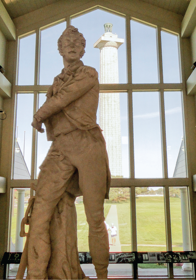 A statue of a man, inside a building; out the windows behind the statue is a tall gray column.