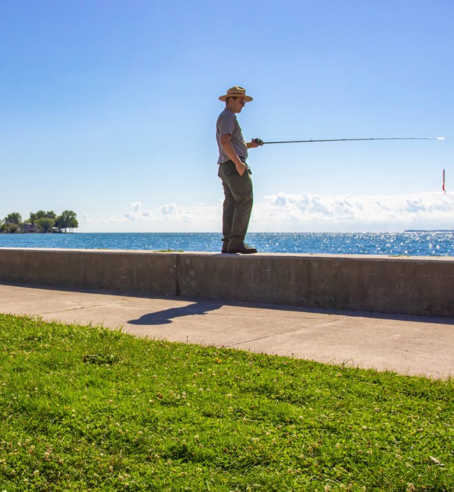 Park Ranger wearing green pants, gray button up shirt, and wearing a straw campaign hat holds fishing pole just before casting. A red lure hangs down 12 inches. Ranger is standing on concrete seawall with the sun shimmering off the blue water of the lake.