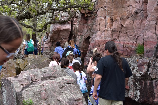 A group of students with a chaperone walk a path along a large stone wall.