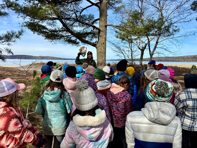 A group of students look at a park ranger holding the iconic Arrowhead. South Bay of Lake Superior is in the background.