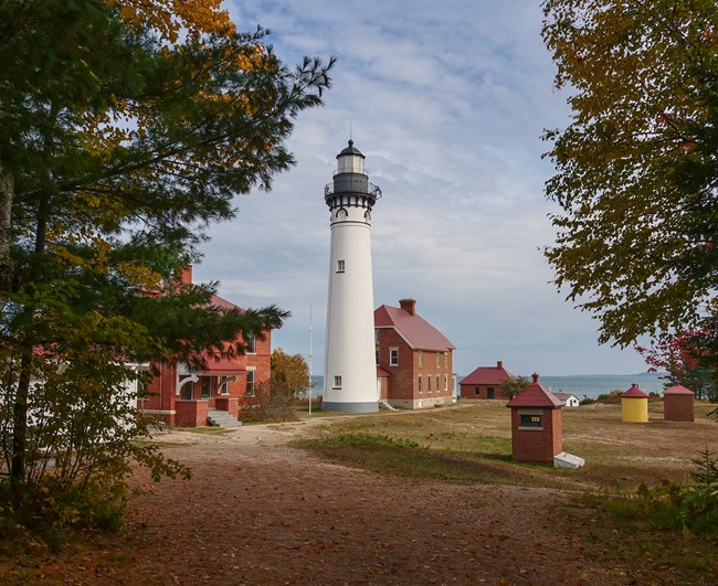 A tall white lighthouse and red brick buildings are seen through a clearing in the trees.