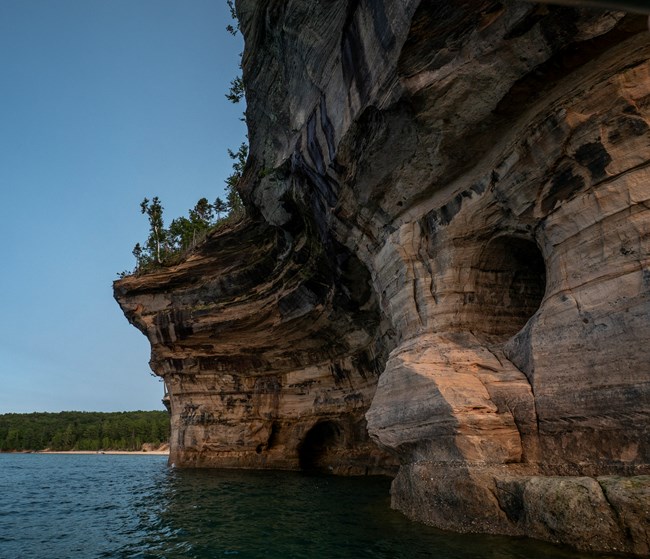Mineral-stained sandstone cliffs rise out of Lake Superior. The portion closest to the water appears to have pebbles stuck all over it. The portion just above that looks cross bedded and has caves.