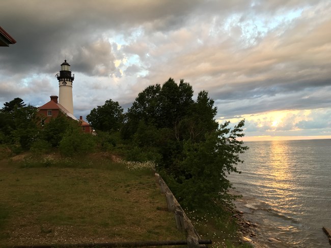 A lighthouse stands above trees along a coastline during a colorful sunset