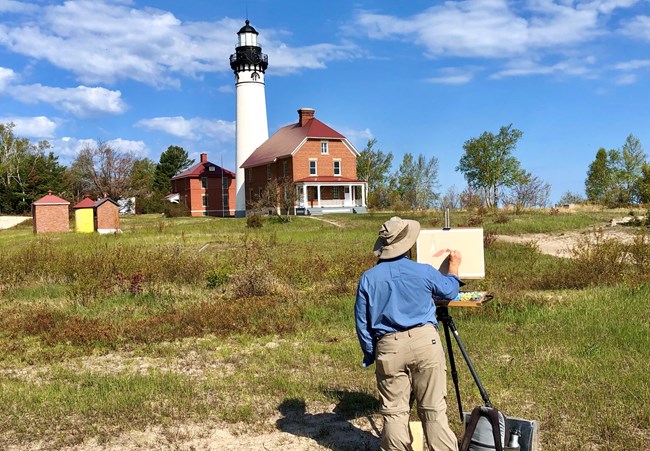 A person with their back to the camera paints a lighthouse, which is in the background