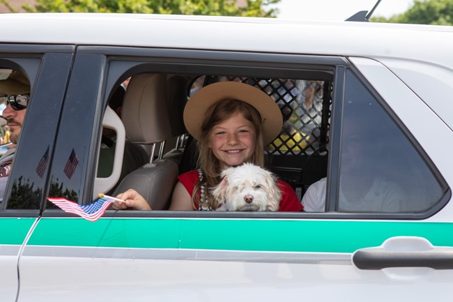 A small white dog sits on the lap of a girl sitting in the back of a park ranger vehicle