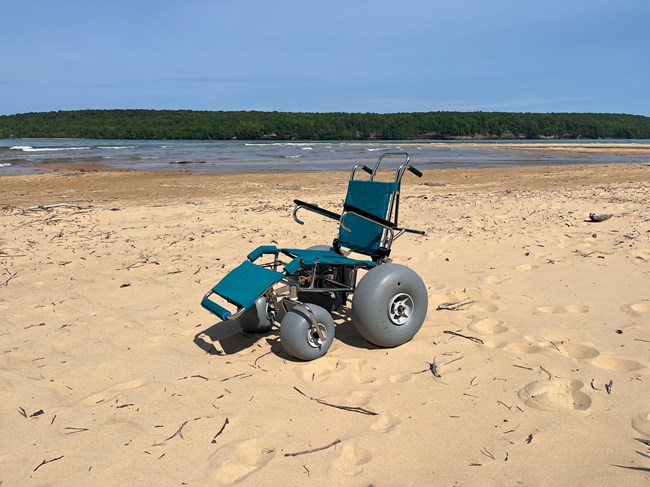 A teal and gray wheelchair with large wheels sits on a beach