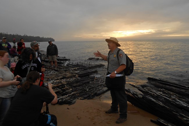 Park ranger stands on a beach next to wooden shipwreck and crowd