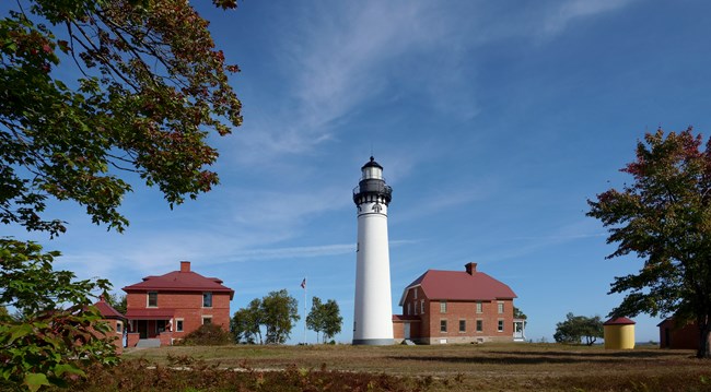 A tall, white lighthouse surrounded by red brick buildings and autumn grass and foliage