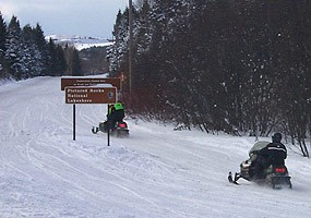 Two snowmobiles travel along County Road H-58 with the Grand Sable Dunes in the background.