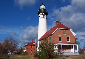 Three Au Sable Light Station buildings including the white-washed towers are silhouetted against a blue sky.