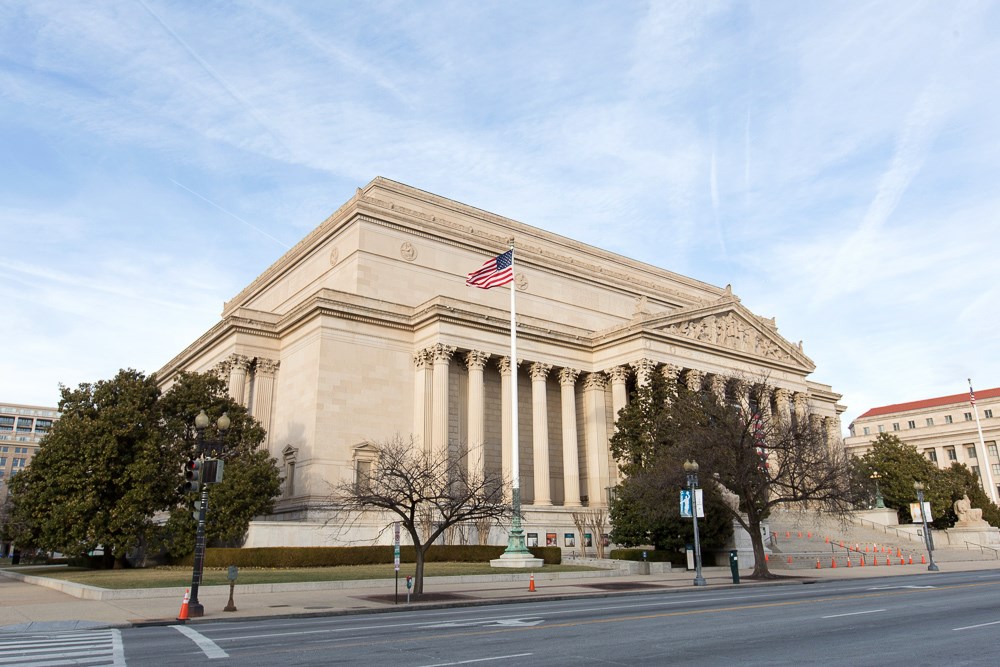 A modern, color photo showing an imposing, classically designed building. It is reminiscent of a Roman temple. The photo has small trees, part of a road, and an American flag on a pole in the foreground of the image.