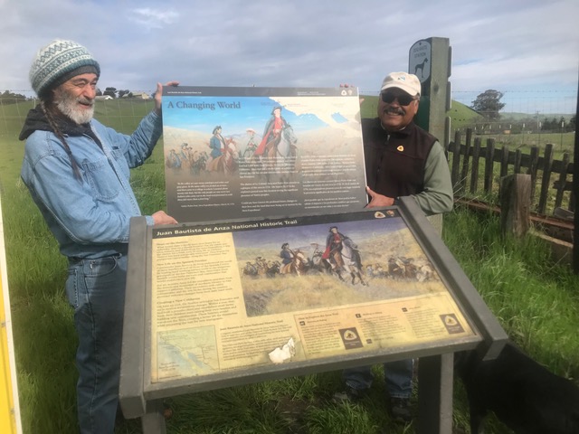 Two volunteers standing behind an old interpretive wayside panel and holding a new panel. 