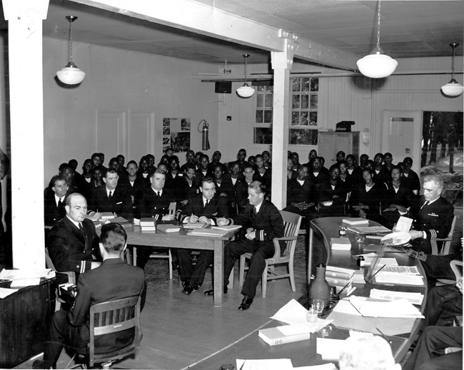 A room full of diverse men during trail proceedings. Some sailors, some suits.