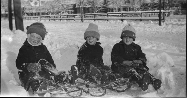 A black-and-white photo of Joe Jr., JFK, and a friend sledding as children.