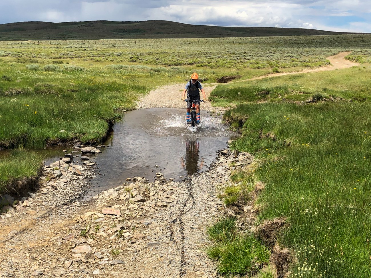 A person on a bike rides through a puddle on a dirt road.