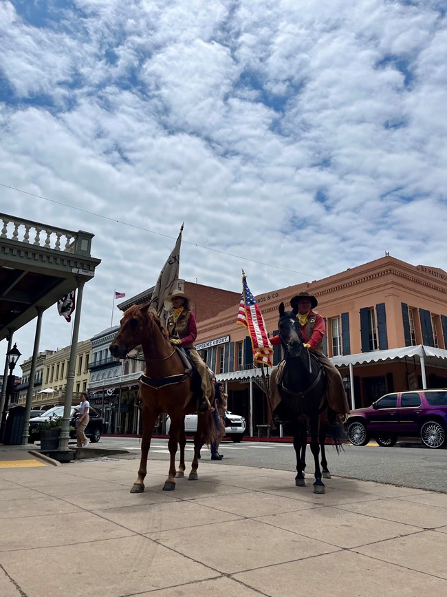 Two people on horseback, with flags, stand in the street lined with historic buildings.