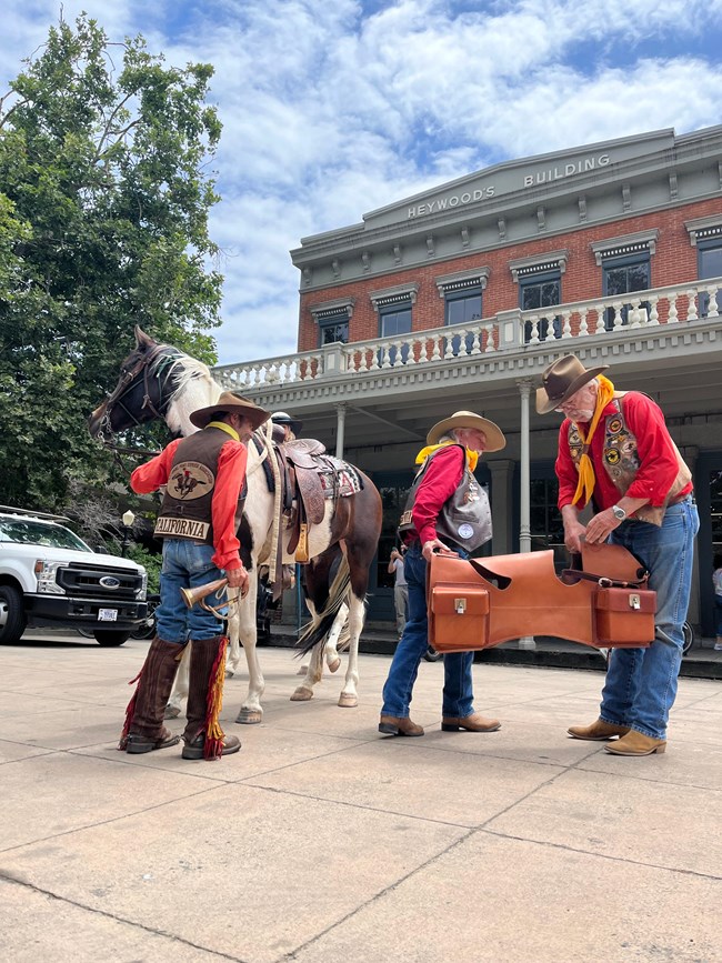 A man hands another man a leather saddlebag, while standing next to a horse.