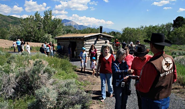 Many people gather around a grey wooden cabin surrounded by green shrubs and trees with mountains in the background.