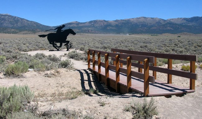 A bridge crosses over a dry drainage with sand and shrubs all around, and mountains in the background with a black steel silhouette of a rider and horse.