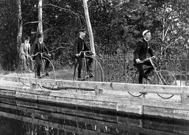 black and white photo of three men riding bikes over a bridge