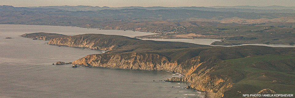 An aerial photo of a narrow grass-covered peninsula with beige cliffs rising above the ocean in the lower left. A narrow bay separates the peninsula from the mainland, which stretches into the distance.