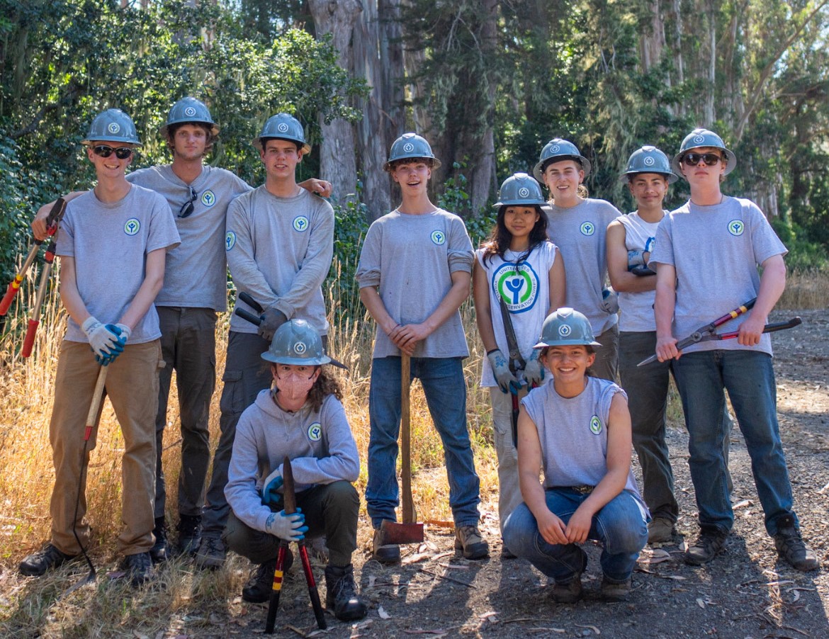 Ten youth in grey t-shirts and various hand tools stand in front of a eucalyptus forest.