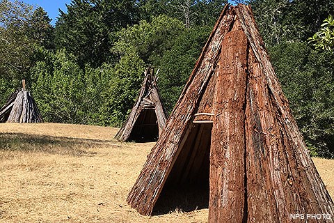 Three ten-foot-tall cone-shaped structures made of redwood-bark sit in a grassy area adjacent to the woods.