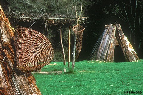 Part of a redwood kotca with a basket (left), a shade structure (center), and a second redwood kotca (right) at Kule Loklo.